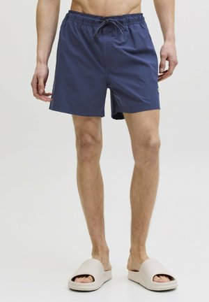 Homme portant un short de bain bleu marine avec cordon de serrage et des sandales beige à enfiler, debout devant un fond clair uni.