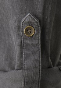 Grått tyg med en slät textur, med en knappstängning i form av en liten cirkel med ett korsmönster, sydd på plats.