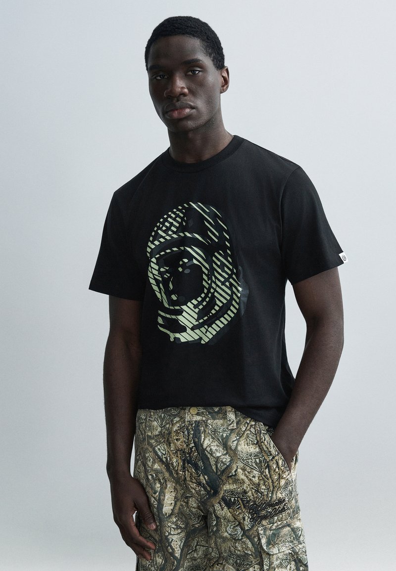 Μαύρο μπλουζάκι με γραφικό ελαφρώς πράσινου αστροναύτη, συνοδευόμενο από μπεζ παντελόνι με σχέδια δέντρων και ζώων.