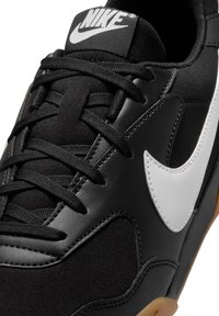 Scarpa sportiva nera realizzata in materiali in pelle e tessuto, con un importante logo swoosh bianco, lacci neri e una suola in gomma naturale.