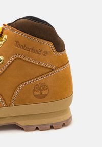 Bota de cuero marrón claro con acentos en marrón, que presenta ojales de metal, costuras blancas y un logo de un árbol. Superficie texturizada y suela resistente visibles.