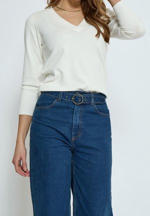 Kobieta ubrana w biały top z długim rękawem i dekoltem w serek, wpuszczony w niebieskie, wysokiej talii dżinsy z paskiem z okrągłą złotą klamrą, z ręką częściowo uniesioną.