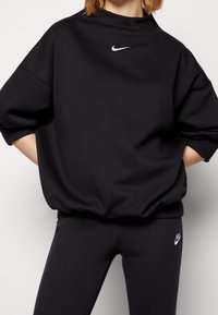 Svart oversize sweatshirt med hög krage, ribbade ärmavslut och liten vit Nike-logotyp på bröstet, matchad med svarta leggings.
