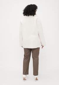 Cream-colored faux fur jacket with a wide collar, long sleeves, and a relaxed fit, paired with brown tailored pants and white high heels.