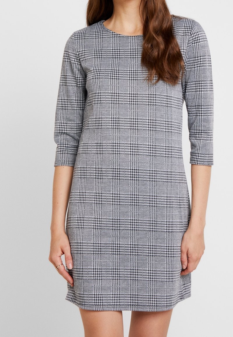 Femme portant une robe à carreaux gris et noirs à manches trois-quarts, debout avec les bras détendus le long du corps.