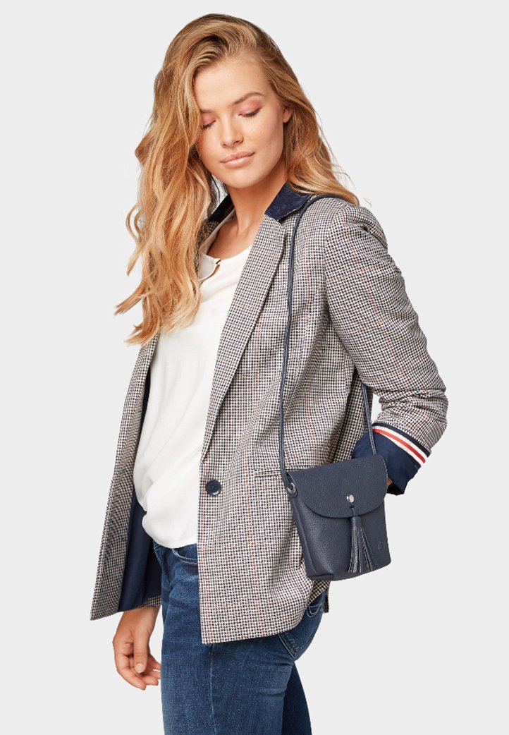 Blazer grigio a scacchi con colletto blu navy, abbinato a una maglietta bianca e jeans blu. Presenta una piccola borsa a tracolla blu navy con dettagli a frange.