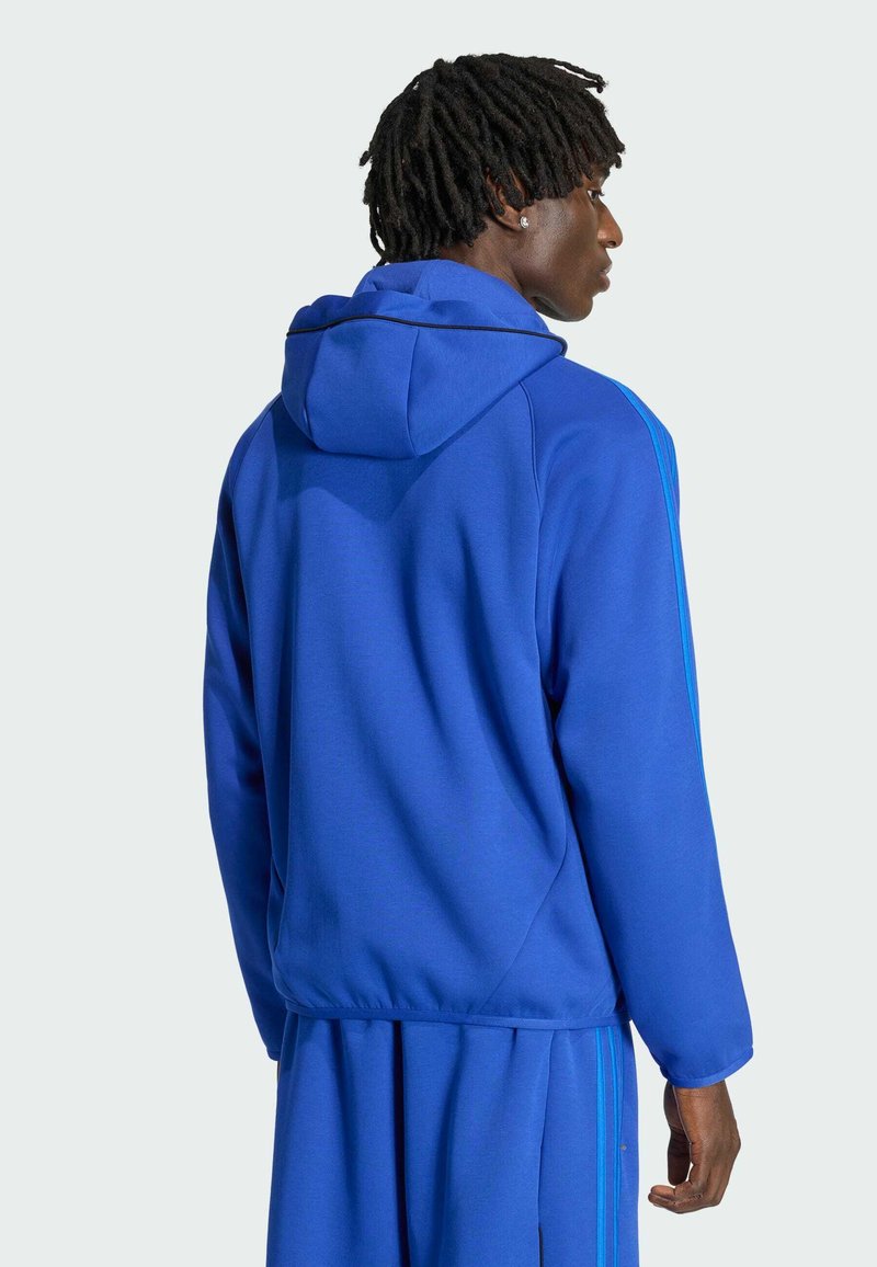 Homme aux cheveux tressés foncés portant une veste à capuche bleue et un pantalon assorti, vu de dos et tourné de côté.