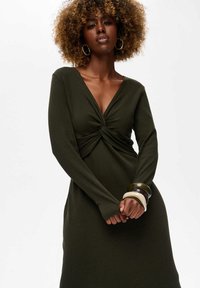 Zielona oliwkowa sukienka z długimi rękawami, dekoltem w serek i skręconym detalem na przodzie. Tekstura prążkowana. Modelka nosi stakowane bransoletki na nadgarstku.