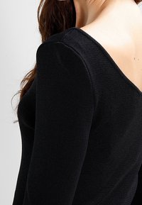Zwarte langharige top met een brede halslijn, strakke pasvorm en textured stof. De achterkant heeft een diepe uitsnijding voor extra detail.