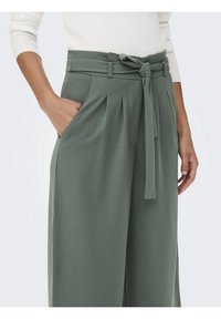 Zelené kalhoty s vysokým pasem, látkovým páskem, záhyby vpředu a bočními kapsami; jemná textura a široký střih nohavic.