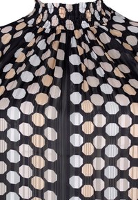 Blouse transparente avec un col smocké haut et un motif d'hexagones noirs, beiges, blancs et gris, avec de douces plis sur l'ensemble.