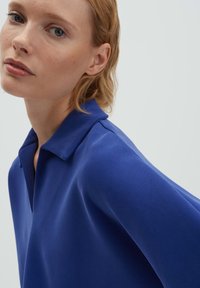 Femme aux cheveux blonds courts portant une chemise bleue à col, regardant légèrement sur le côté sur un fond clair et uni.