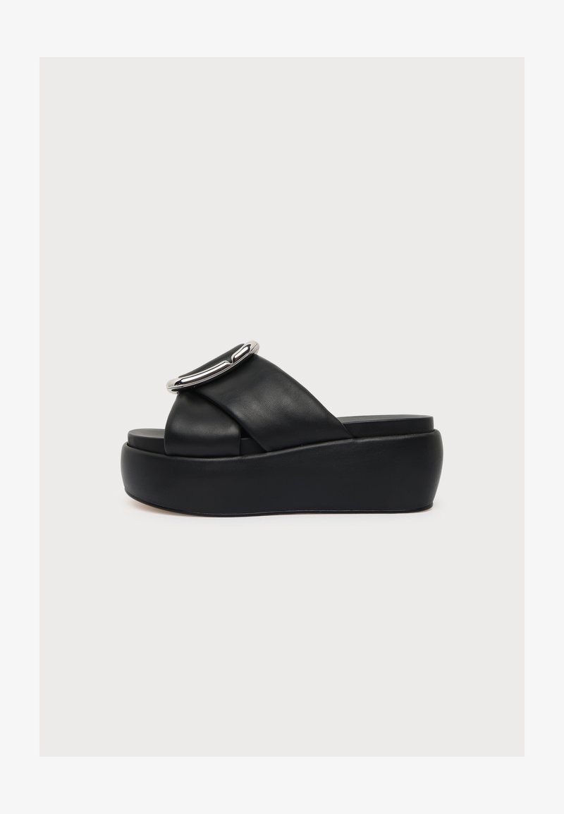 Sandale noire à plateforme avec de larges brides en cuir croisées et une grande boucle argentée sur fond blanc.