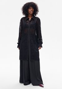 Czarna, długa koszula z koronki z zapięciem na guziki z przodu, posiadająca długie rękawy i podszewkę w jednolitym czarnym kolorze. Teksturowana wzorami kwiatowymi.