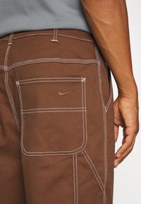 Bruna tygbyxor med vit sömnad, som har en bakficka och Nike-logga i brunt broderi. Högkvalitativ denimtextur är synlig.