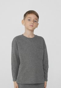 Světle šedá dlouhá košile s dlouhým rukávem vyrobená z měkké látky, s kulatým výstřihem a jednou náprsní kapsou. Jednoduchý, volný střih.