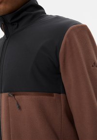 Giacca in pile con parte superiore nera e parte inferiore marrone, dotata di una tasca con zip e tessuto testurizzato per calore e comfort.