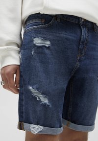 Short en jean bleu foncé avec déchirures effilochées et revers retroussés. Présente des surpiqûres contrastantes et un design classique à cinq poches.