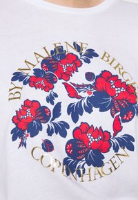 Camiseta de algodón blanca con un estampado floral circular en rojo y azul que incluye el texto dorado "By Malene Birger Copenhagen" alrededor del diseño.
