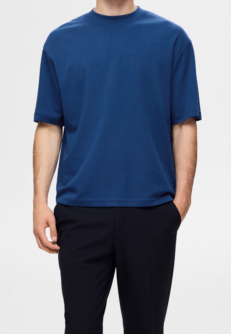 T-shirt bleu à manches courtes en doux tissu de coton, coupe décontractée, encolure ronde et détails minimaux. Porté avec un pantalon sombre.