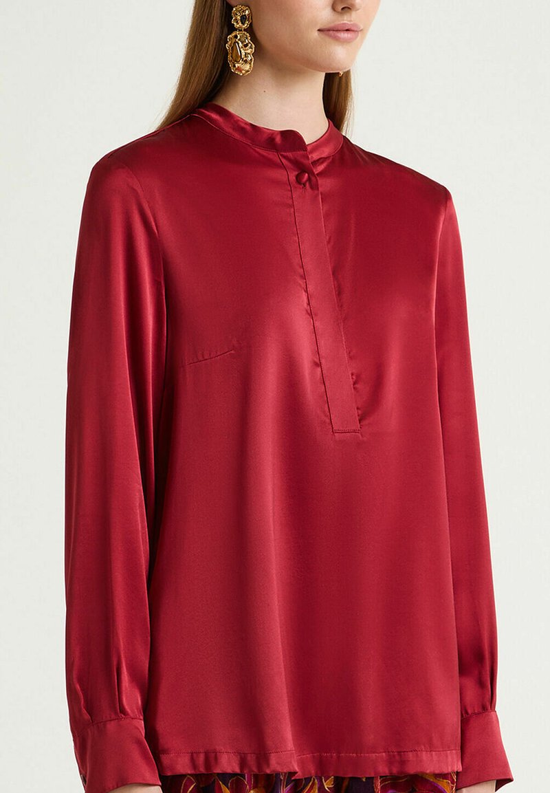Rote Satinbluse mit langen Ärmeln, hohem Kragen, vorderer Knopfleiste und einer aufgesetzten Tasche auf der linken Seite. Glatte Textur mit lockerem Sitz.