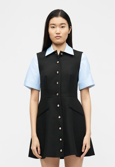 Γυναίκα που φοράει μαύρο αμάνικο φόρεμα με χρυσά κουμπιά πάνω από ανοιχτό μπλε πουκάμισο με κοντά μανίκια και γιακά, στέκεται μπροστά σε μονόχρωμο φόντο.