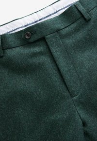 Green wool trousers with a textured finish, featuring a button closure, flat front design, and a white inner waistband with blue stitching.