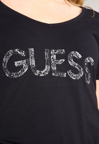 Camiseta de algodón negra con escote en V y la palabra "GUESS" adornada con lentejuelas plateadas y detalles de encaje.