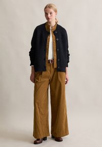 Cardigan nero a maglia con grandi bottoni, abbinato a una maglietta in corduroy senape e pantaloni a gamba larga, stivali marroni con tacco, sfondo semplice.