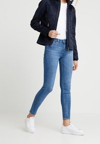 Marinblå fleecejacka, vit t-shirt, blå smala jeans och vita sneakers. Outfiten har en dragkedja framtill och en figurnära design.