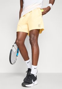 Κίτρινες αθλητικές βερμούδες με δύο πλαϊνές τσέπες και λεπτομέρεια λογότυπου. Συνδυασμένες με μαύρα αθλητικά παπούτσια και λευκές κάλτσες. Πρακτική ρακέτα τένις στο χέρι.