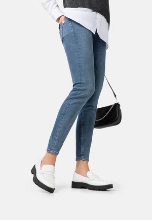 Mocassins blancs avec une surface texturée et de grosses semelles en caoutchouc noir, associés à un jean skinny bleu et un sac à bandoulière noir, avec une chaîne en argent.