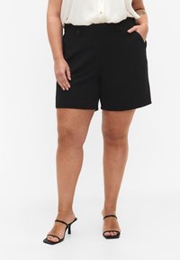 Kvinna i svarta högmidjade shorts, krämfärgad ärmlös blus och svarta klacksandaler med remmar, står med ena handen i fickan.