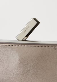Calvin Klein lommebok i lys taupe skinn med teksturert overflate. Har en skinnende sølvfarget metalldeksel med merkevarenavnet gravert.