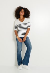 Suéter de rayas blancas y marinas con escote en V y mangas cortas, combinado con jeans acampanados azules y zapatillas blancas. Presenta un logo en la manga.