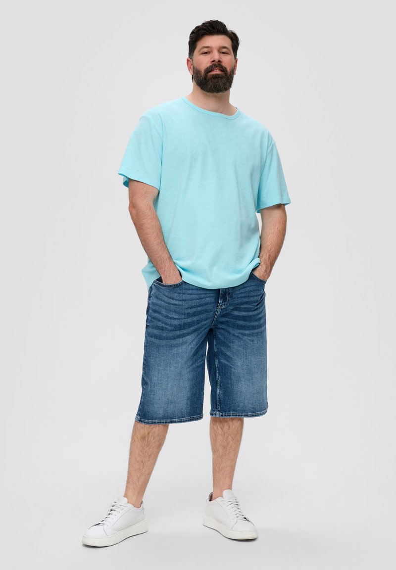 Lichtblauw T-shirt met korte mouwen, denim shorts en witte sneakers. Model staat met handen in de zakken tegen een grijze achtergrond.