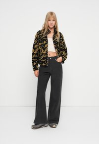 Chaqueta de forro polar con patrón de tie-dye negro y dorado, diseño cropped. Combinada con pantalones anchos de talle alto en negro y zapatillas grises.