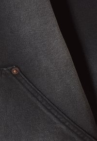 Prim-plan al unui material textil negru, care arată o suprafață texturată, detalii de cusături și un nasture de cupru pe un buzunar. Fără modele vizibile.
