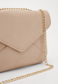 Béžová kabelka z ekologické kůže přes rameno s klopovým uzávěrem, zlatým řetízkovým popruhem a šitými detaily. Hladký povrch s elegantním designem.