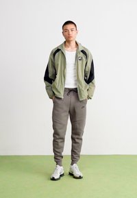 Wiatroszczelna kurtka Nike w jasnym zielonym kolorze z czarnymi akcentami, zestawiona z szarymi spodniami dresowymi. Białe sneakersy dopełniają sportowy wygląd. Gładka faktura, swobodny fason.