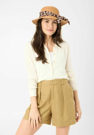 Femme portant un chapeau de paille beige avec un nœud à motif léopard, un cardigan crème boutonné, un haut blanc et un short beige taille haute, souriant légèrement.