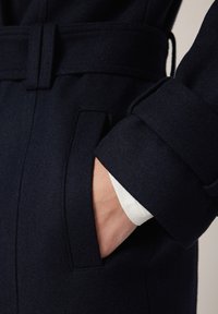 Szczegół marynarskiego wełnianego płaszcza z dopasowanym krojem, boczną kieszenią i mankietem z kontrastowym białym wykończeniem. Powierzchnia jest gładka.