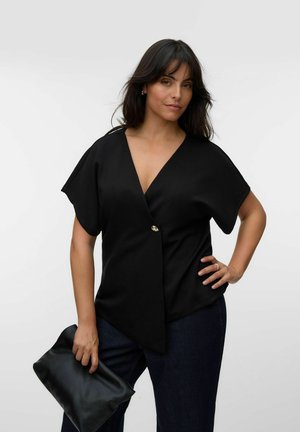 Femeie cu păr închis la culoare, purtând un top negru cu decolteu în V și pantaloni închiși la culoare, ținând o poșetă neagră, pozând cu o mână în șold pe fundal alb.