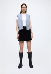 Veste tricotée bleu clair avec boutons, t-shirt blanc, jupe en velours côtelé noire et bottes hautes noires. Sur un fond uni.