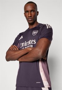 T-shirt de sport violet foncé à manches courtes, avec un logo "Emirates" blanc, des accents latéraux en dégradé rose et un col rond.