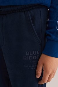 Jūras spilventiņi ar sānu kabatu, elastīgu jostasvietu un iegrieztu logotipu. Materiāls izskatās mīksts ar gludu tekstūru. Redzama zilā garā piedurkņu kreklā.