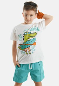 Hvid t-shirt med et legende krokodillegrafik, parret med teal shorts. Funktioner inkluderer lyse farver, en afslappet pasform og et strandtema.