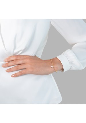 Bracelet en or avec une chaîne délicate et un charm en forme de cœur rose, porté sur une main contre un fond clair, associé à un blouse blanche.