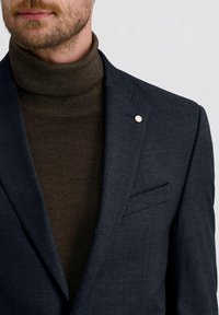 Marineblazer met een getextureerde, donkerbruine turtleneck. De blazer heeft een elegante uitstraling en een metallic accent op de revers.