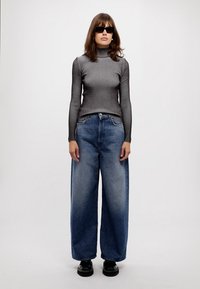 Un col roulé gris à côtes, un jean en denim bleu clair à jambes larges, des chaussures noires imposantes, avec des lunettes de soleil sombres et des accessoires minimalistes.